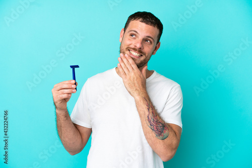 Brazilian man shaving his beard isolated on blue background looking up while smiling