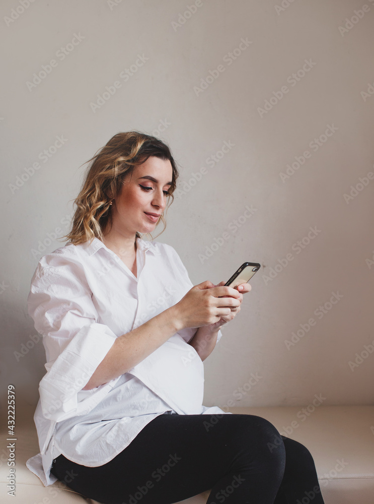 A pregnant woman keeps and uses her smartphone at home. Close-up. A young pregnant woman holds her stomach and looks at her mobile smartphone.