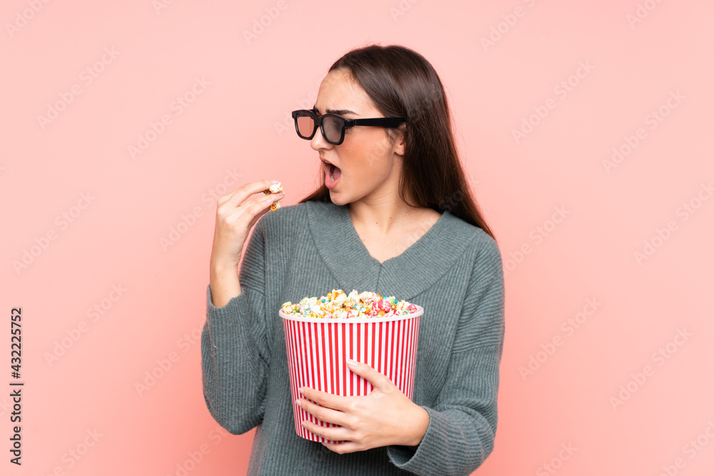 Young caucasian woman isolated on pink background with 3d glasses and holding a big bucket of popcorns while looking side Young caucasian woman isolated on pink background with 3d glasses and holding a big bucket of popcorns while looking side