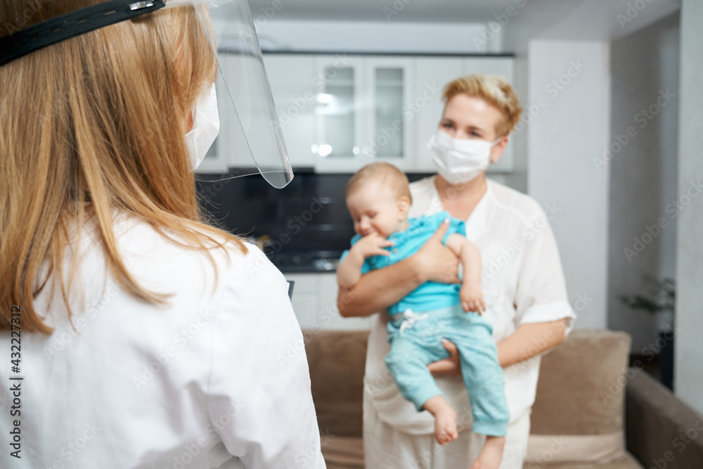 View from shoulder in medical uniform and mask talking with young woman that holding cute boy on hands. Health care at home. View from shoulder in medical uniform and mask talking with young woman that holding cute boy on hands. Health care at home.