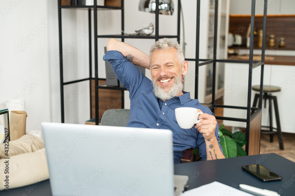 Fototapeta premium Cheerful mature male employee enjoys morning coffee at the workplace in the office, sits in relaxed pose with feet on the table, satisfied middle aged man in smart casual wear takes a break