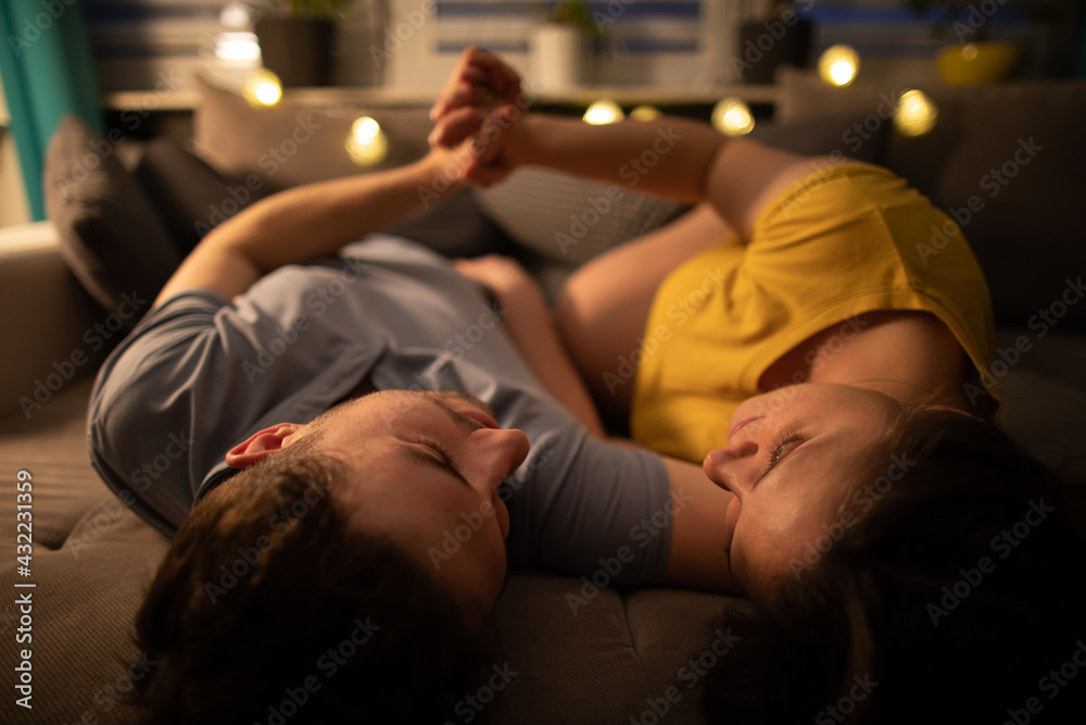 A married couple lying in bed in the evening holding hands. The woman in advanced pregnancy.