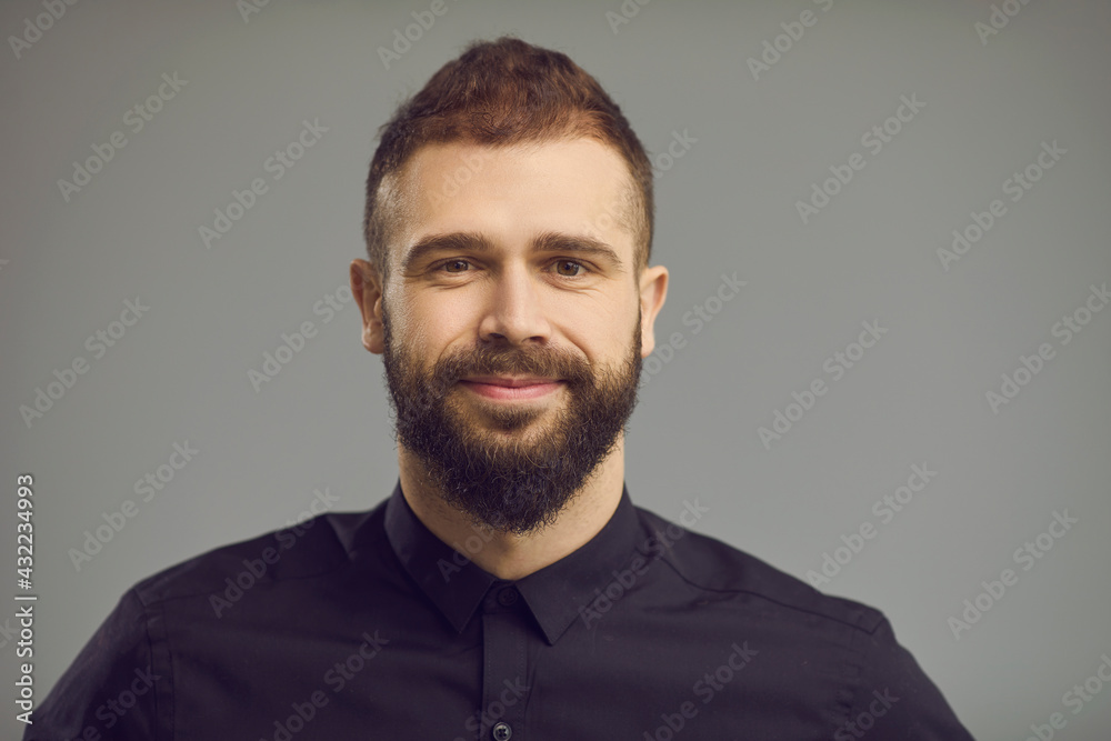 Handsome people emotion and expression concept. Attractive caucasian hipster bearded male model with friendly smile on face wearing stylish shirt standing over gray studio backgound. Headshot portrait