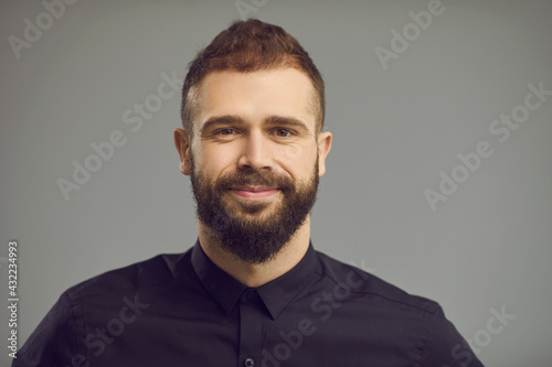 Handsome people emotion and expression concept. Attractive caucasian hipster bearded male model with friendly smile on face wearing stylish shirt standing over gray studio backgound. Headshot portrait