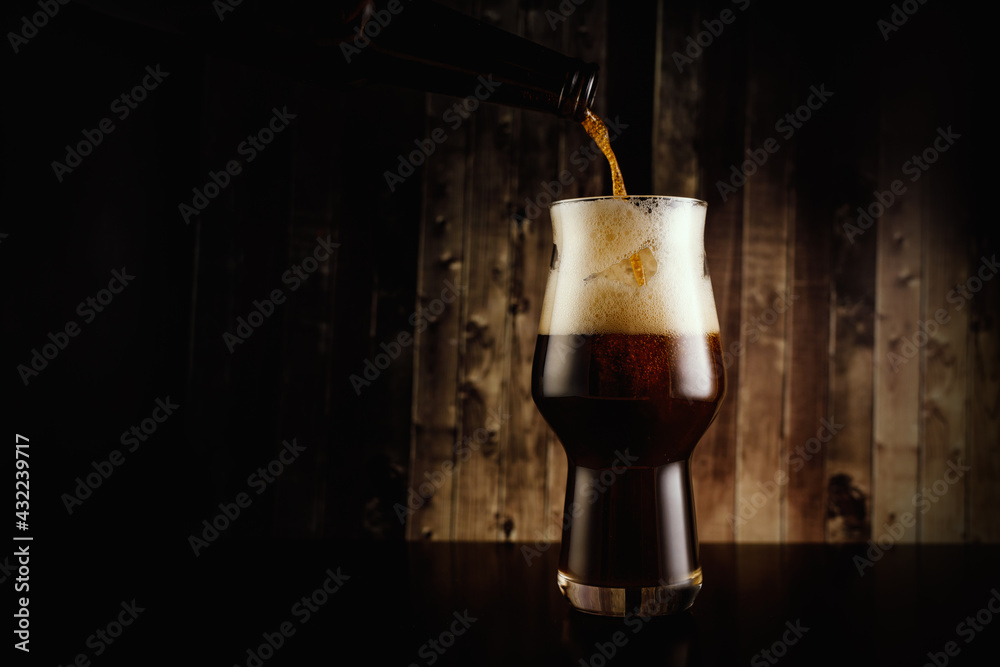Pouring dark beer into the glass from the bottle on wooden background. - obrazy, fototapety, plakaty 