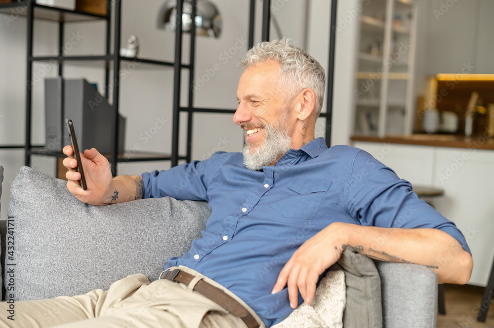 Contemporary charismatic mature man in casual shirt using smartphone lying down on the couch at home, bearded handsome male spends leisure online, watching videos on the phone, scrolling news feed Contemporary charismatic mature man in casual shirt using smartphone lying down on the couch at home, bearded handsome male spends leisure online, watching videos on the phone, scrolling news feed