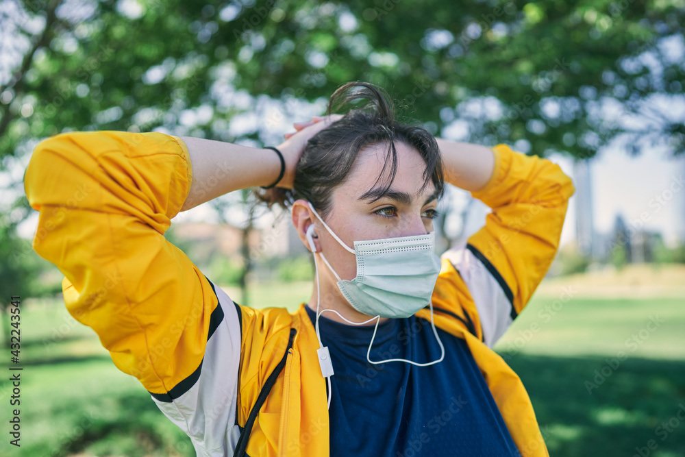 Naklejka premium A beautiful white woman with brown hair and a sports suit with a yellow jacket prepares for exercise and exercises her muscles to avoid cramps in a park near the city wearing a Covid 19 mask 