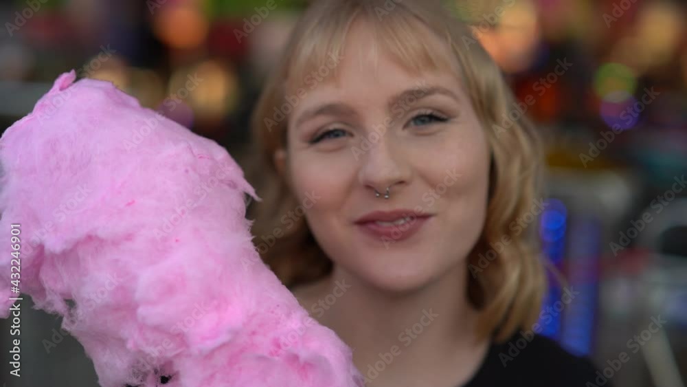 Primer plano de chica rubia con ojos azules guapa mirando a cámara y comiendo algodón de azúcar rosa en zona de atracciones de una feria