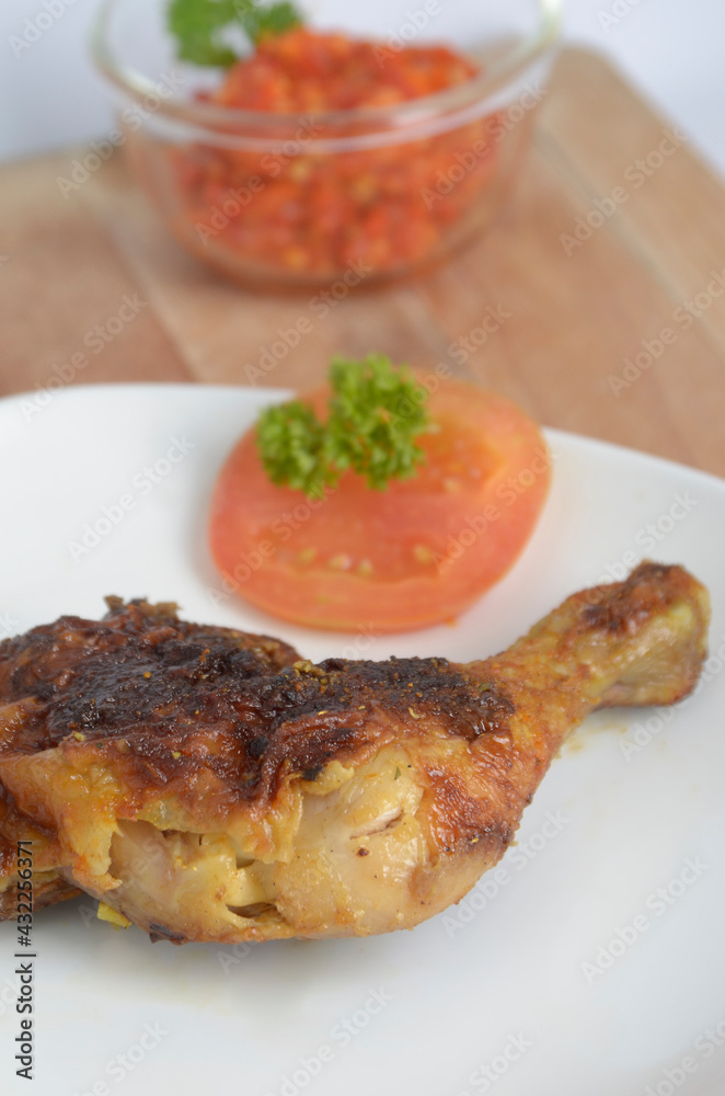 grilled chicken thigh serve with slice tomato as garlic in white plate, and sambal or sambel ( Chili sauce ) in ceramic bowl.