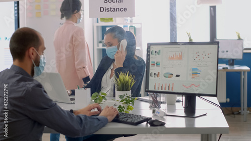 Coworkers with face masks sitting at desk in new normal company office working on laptop compuer, talking on phone during coronavirus pandemic. Team mantain social distancing to avoid virus disease photo