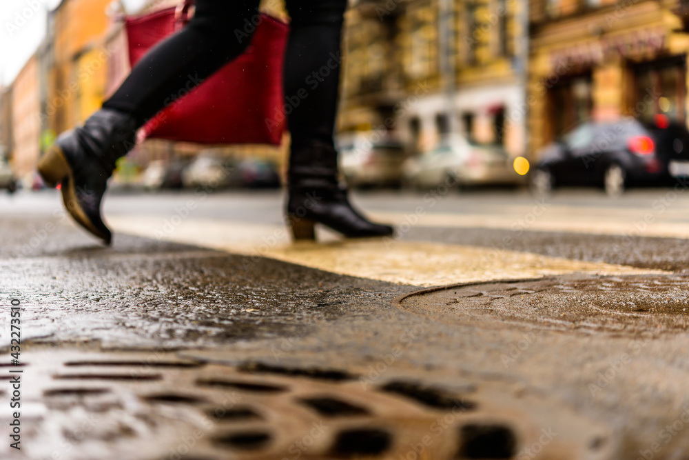 Rainy day in the big city, the woman with a red bag crosses the road on a pedestrian crossing. Close up view of a hatches at the level of the asphalt