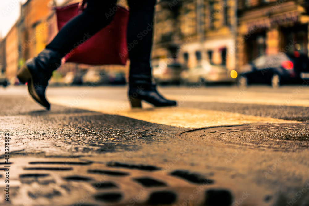 Fototapeta premium Rainy day in the big city, the woman with a red bag crosses the road on a pedestrian crossing. Close up view of a hatches at the level of the asphalt
