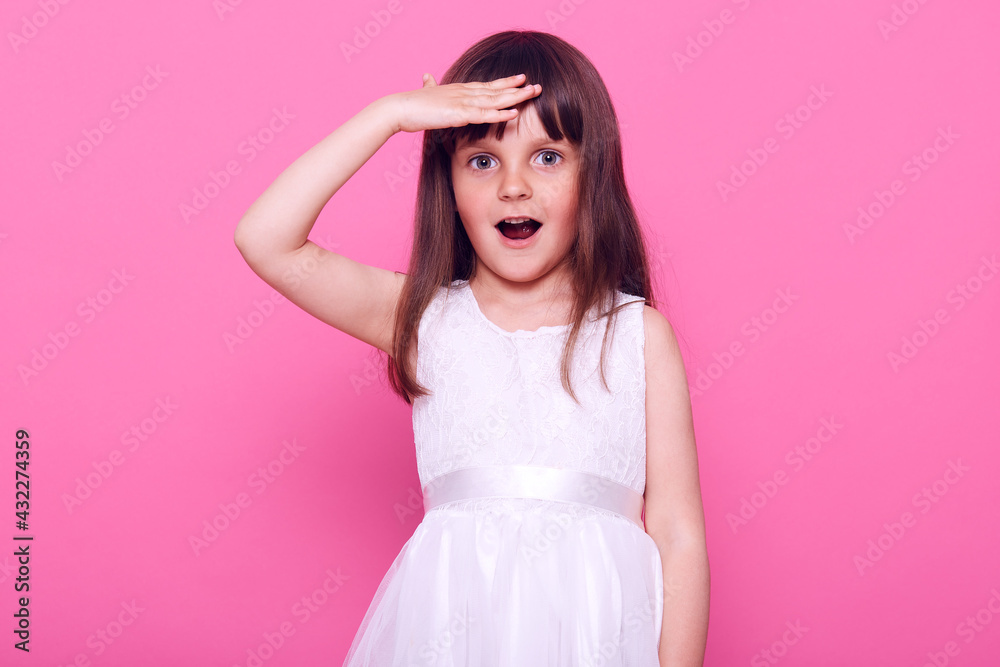 Astonished little girl wearing stylish white dress looking far away with opened mouth and surprised expression, has pleasant surprise, isolated over pink background.