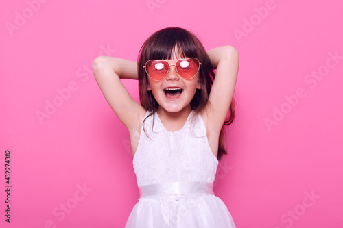 Fotografie Happy small female kid wearing elegant white dress and heart-shaped glasses looks at camera with excited expressing, being glad of great event, raising hands, isolated over pink background