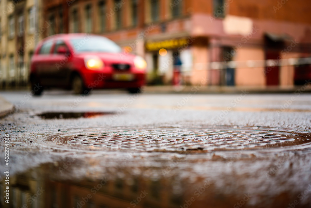Rainy day in the big city, the red car is at the crossroads. Close up view of a hatch at the level of the asphalt