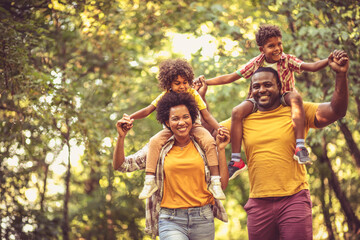 African American family walking trough park. Parents carrying children on piggyback.