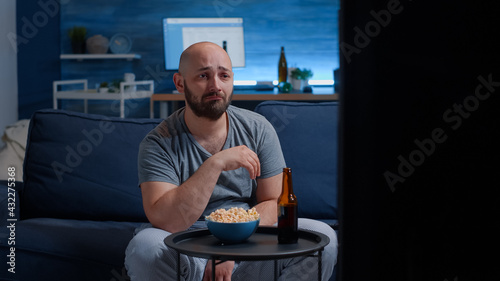 Slika na platnu Sad guy watching drama movie on TV crying wiping eyes sitting on couch alone at night looking at television, eating popcorn