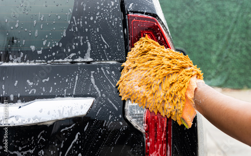 The young man washed his car with foamy cleaning foam. He was wearing gloves for washing the car. He wiped the car's tail lights and body.