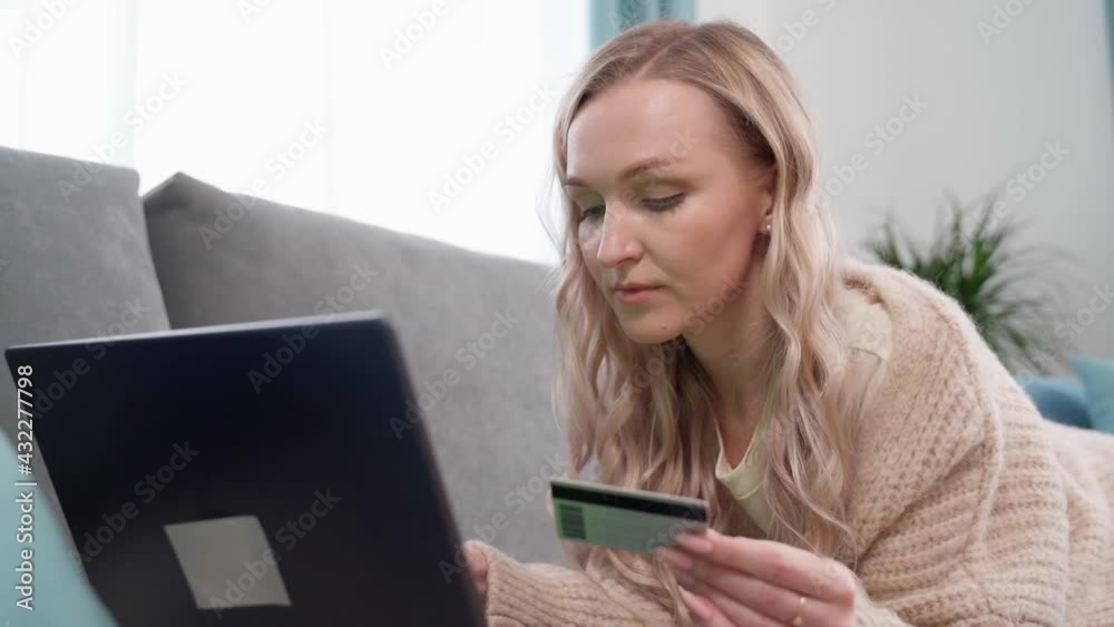 Beautiful blonde lying on the couch makes online payments with a bank card using a laptop at home. Online shopping. Beautiful blonde lying on the couch makes online payments with a bank card using a laptop at home. Online shopping.