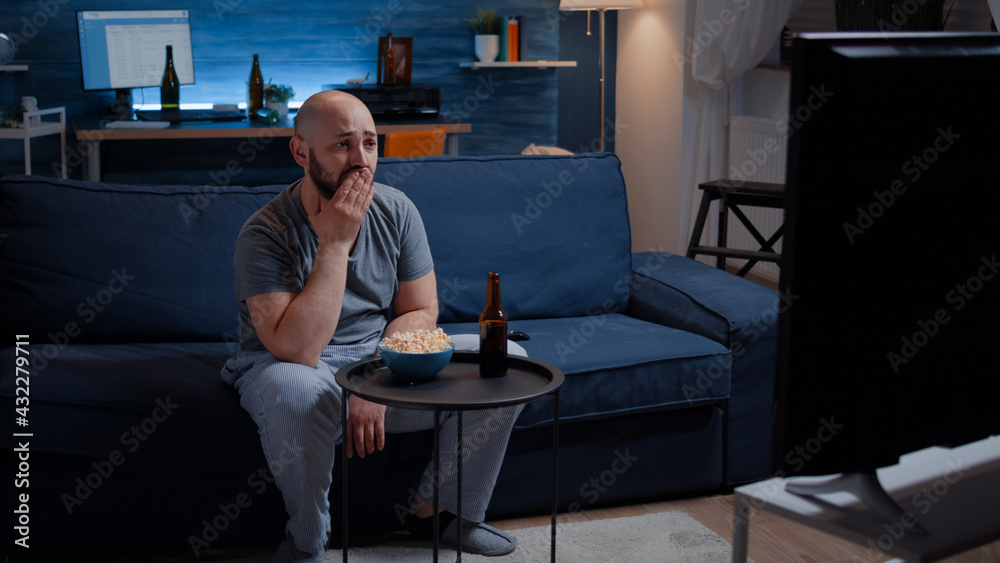 Fotka „Focused man looking at drama movie, crying sitting on sofa eating popcorn late night