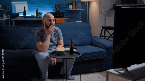 Fotografija Focused man looking at drama movie, crying sitting on sofa eating popcorn late night