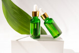 Green glass cosmetic bottles with a dropper on a white podium with tropical leaves. Natural cosmetics concept, natural essential oil and skin care products