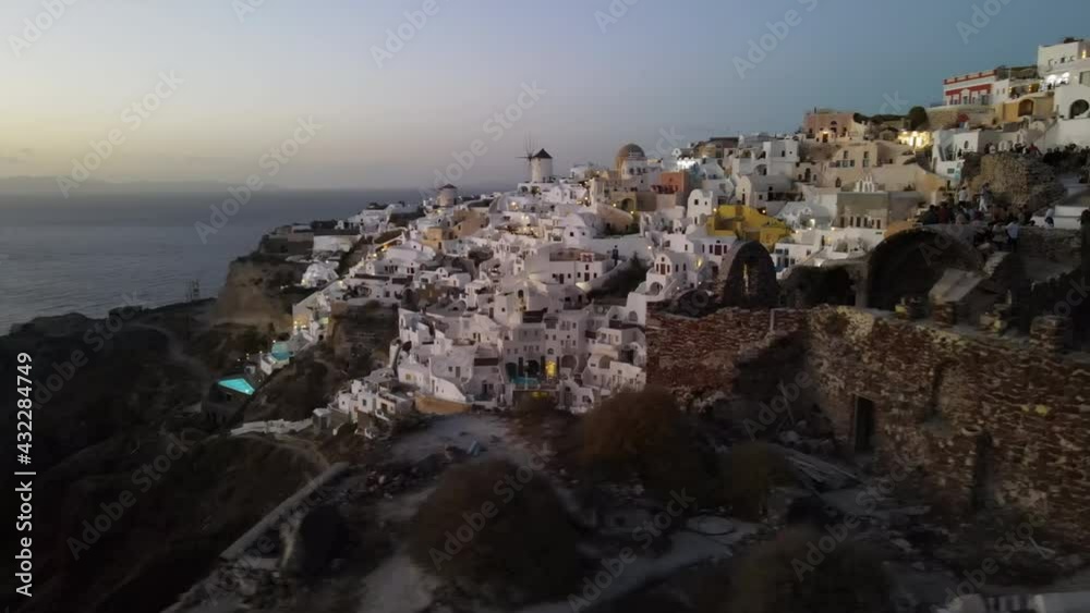 Aerial Panning Shot Of Tourists In City On Mountain Against Sky, Drone Flying Over People On Structure At Sunset - Santorini, Greece Aerial Panning Shot Of Tourists In City On Mountain Against Sky, Drone Flying Over People On Structure At Sunset - Santorini, Greece