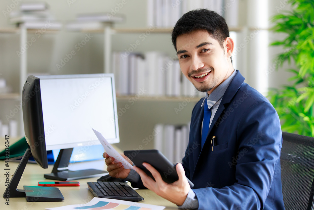 Fototapeta premium Successful young Asian businessman smiles happily while seeing good news on tablet in office. Business and success concept.