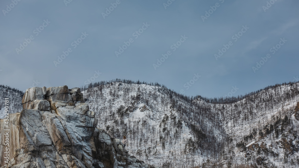 A flat-topped rock devoid of vegetation. Cracks on the stones. Background - wooded snow-covered hills, blue sky. Close-up. Baikal