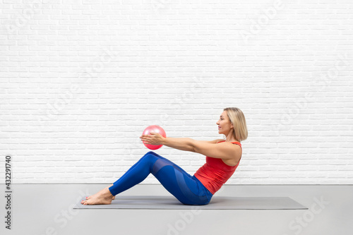 Attractive fit caucasian woman in sportswear sits on the mat and practice roll up drill with pink mini-ball in hands. Pilates indoor with props. photo