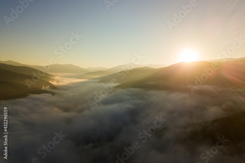 Aerial view of vibrant sunset over white dense foggy clouds with distant dark silhouettes of mountains on horizon.