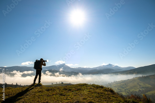 Silhouette of a backpacker photographer taking pictures of morning landscape in autumn mountains with digital camera.