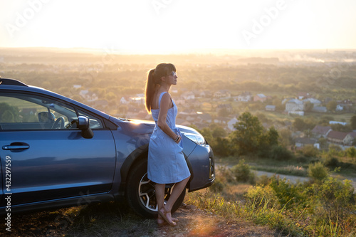 Yong happy female driver resting near her car enjoying sunset view of summer nature. Travel destinations and recreation concept. © bilanol