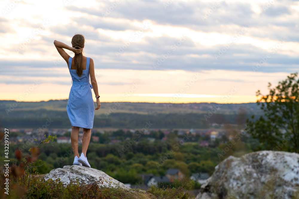 A young woman in summer dress standing outdoors enjoying view of bright yellow sunset.