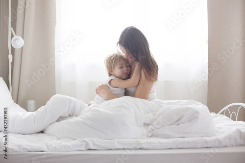 Beautiful blond child and his mother, playing at home in the morning in bed, smiling, laughing and having fun