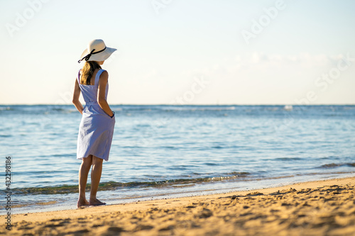 Young woman in straw hat and a dress walking alone on empty sand beach at sea shore. Lonely tourist girl looking at horizon over calm ocean surface on vacation trip.