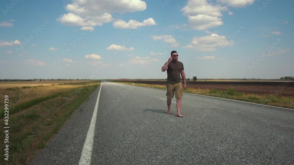 Bearded Young Man Wearing Glasses, T-shirt and Shorts Walking Barefoot on a Deserted Automobile Road Between Fields on a Hot Sunny Day. Hipster Hicker's Traveling Concept Bearded Young Man Wearing Glasses, T-shirt and Shorts Walking Barefoot on a Deserted Automobile Road Between Fields on a Hot Sunny Day. Hipster Hicker's Traveling Concept