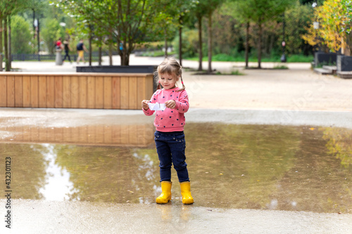 a little girl in a pink sweater and rubber boots launches a paper boat in a large puddle © Елена Гурова