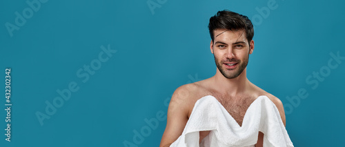 Studio portrait of cheerful brunette guy using a white towel to wipe himself after shaving and taking a morning shower, smiling at camera, posing isolated over blue background