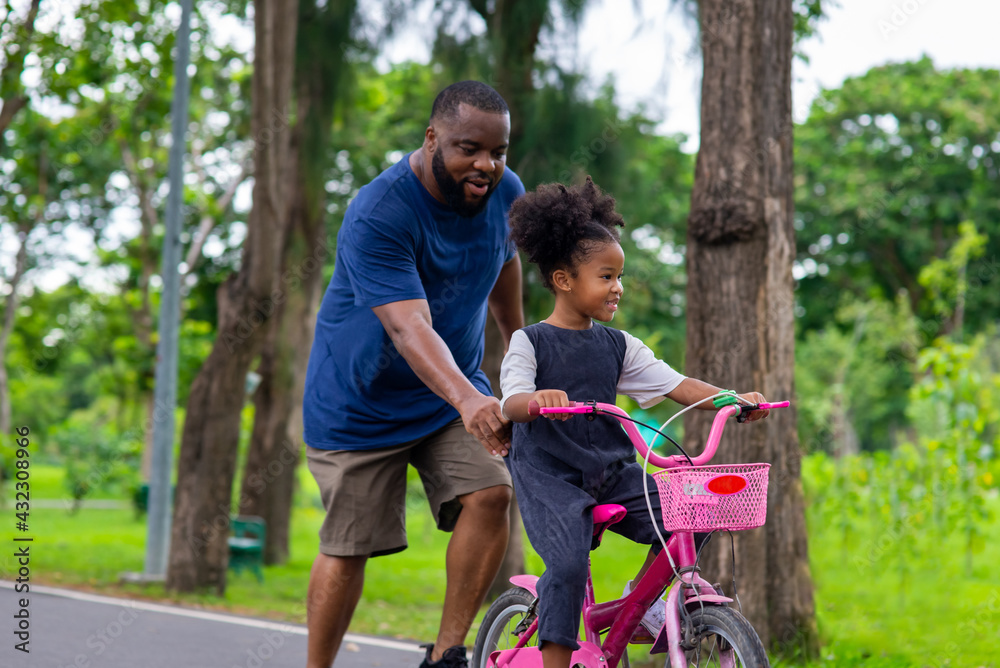 Fototapeta premium Happy mixed race family in park. African father teaching little daughter to ride a bicycle in the park. Dad and child girl kid having fun together outdoor lifestyle activity in weekend vacation.