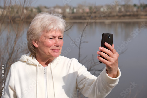 Portrait of happy blonde senior woman holding camera and taking selfie. Selective focus, Focus on phone