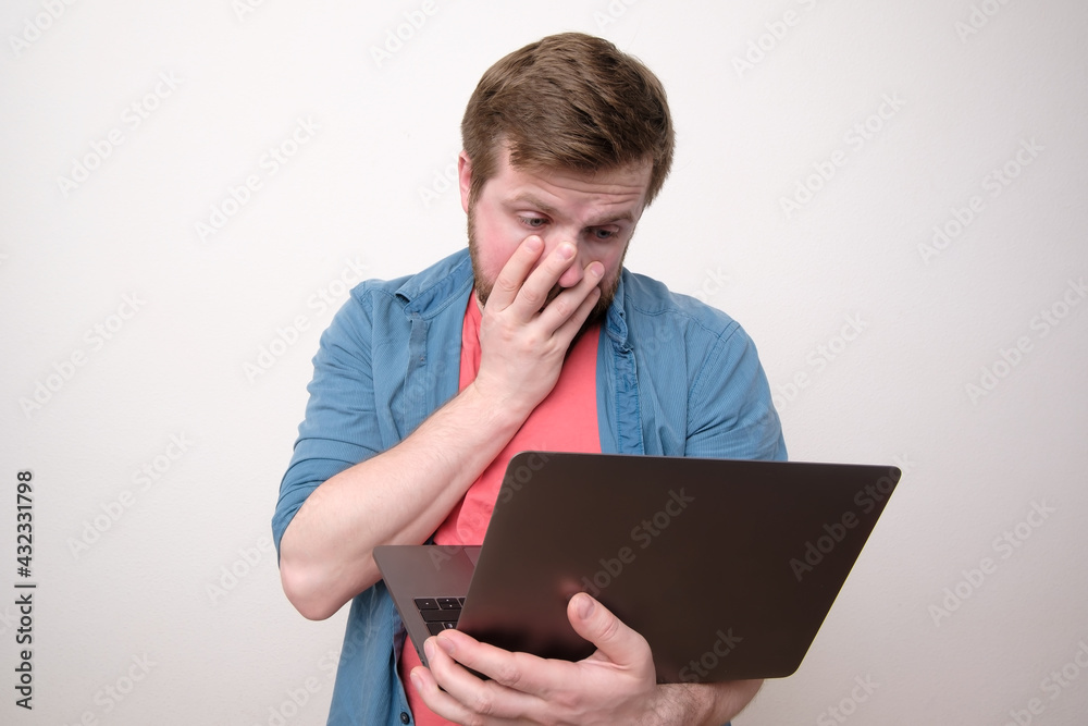 Upset man holds a laptop in hands and looks at the screen in bewilderment, covering part of face with hand. Remote work or education. Upset man holds a laptop in hands and looks at the screen in bewilderment, covering part of face with hand. Remote work or education.