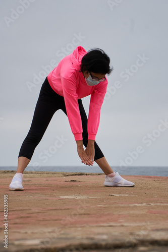 Woman with mask performs stretching exercises in front of the sea. New normality.