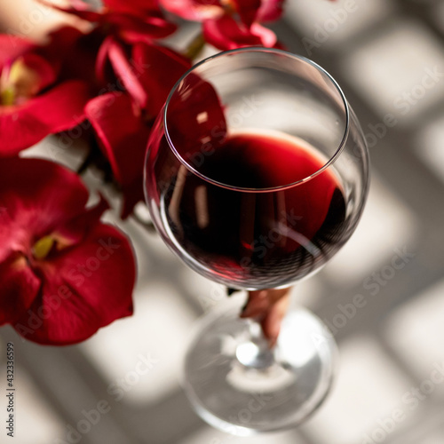 Glass of red wine and red orchid flowers on wooden table with sun glare. Alcoholic drink. Close up shot  soft focus.