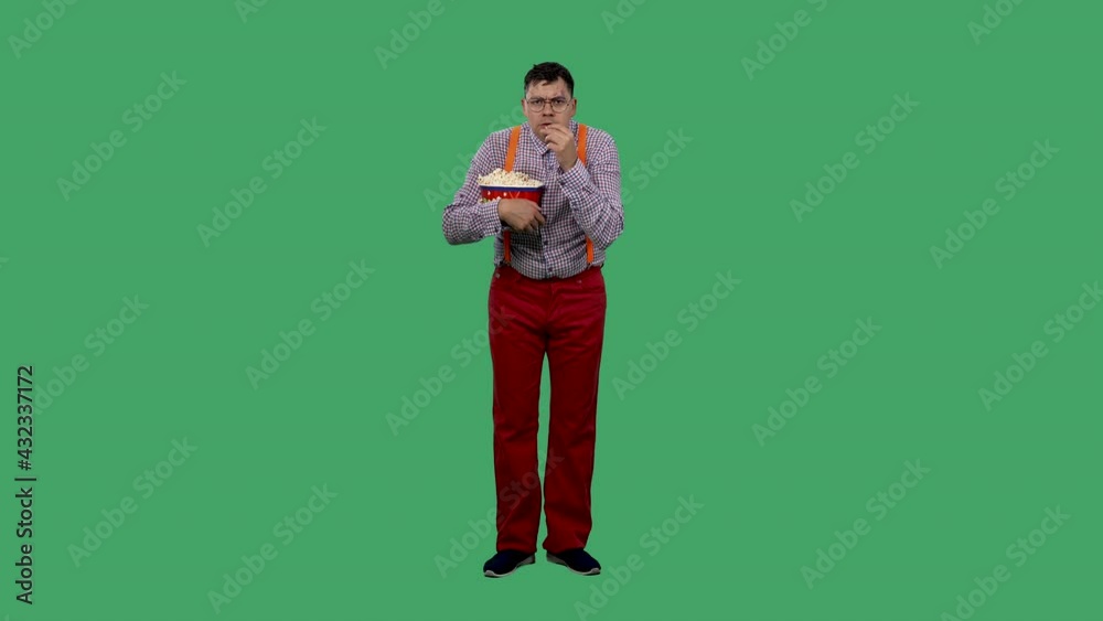 A man watches a movie carefully and eats popcorn from a large bucket. Portrait of a man in glasses, in a shirt with orange suspenders in the studio on a green screen. Slow motion. A man watches a movie carefully and eats popcorn from a large bucket. Portrait of a man in glasses, in a shirt with orange suspenders in the studio on a green screen. Slow motion.