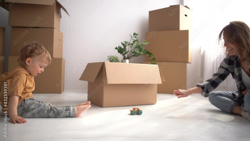 Young mother plays with a child sitting on the floor against the background of cardboard boxes. The family moves to a new apartment, mom and son have a fan during the relocation Young mother plays with a child sitting on the floor against the background of cardboard boxes. The family moves to a new apartment, mom and son have a fan during the relocation