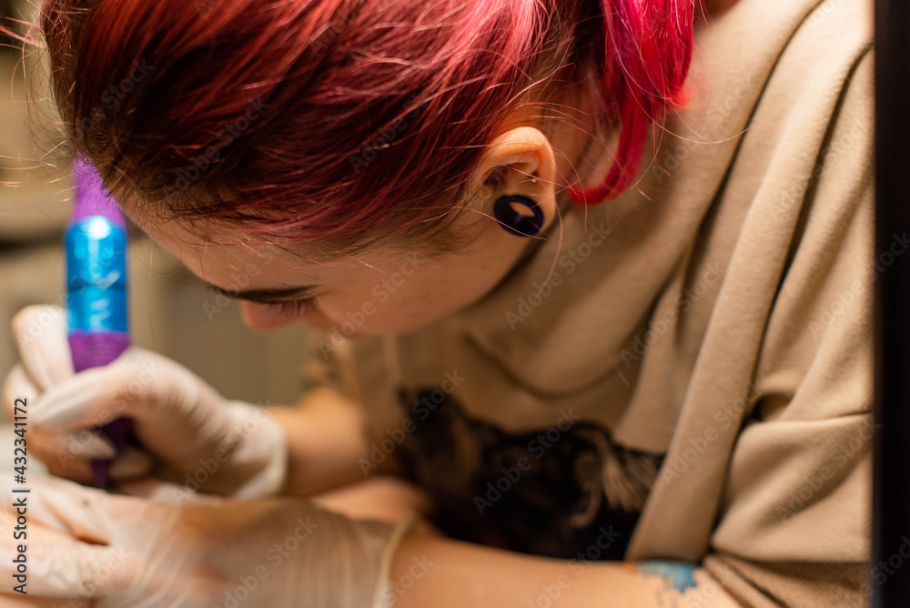 Fototapeta premium Charming girl with pink hair and tattoos stuffs a tattoo on a woman's hand in a tattoo parlor, creative small business and tattoo master. Hipster woman in gloves draws a tattoo