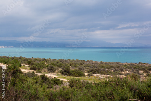 Landscape of Cabo de las Huertas, in Alicante, with vegetation in the foreground, the calm Mediterranean Sea and mountains on the horizon on a cloudy day.