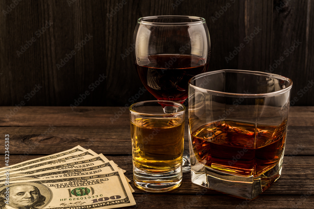 Three glasses with brandy, tequila and red wine with money on an old wooden table. Angle view Three glasses with brandy, tequila and red wine with money on an old wooden table. Angle view