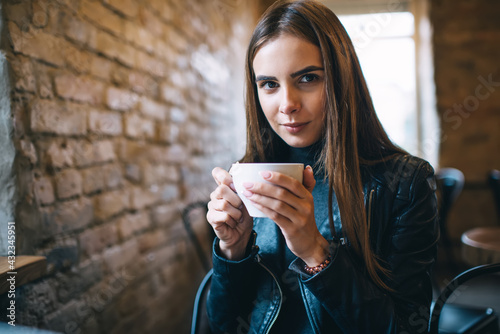 Half length portrait of attractive female customer with cappuccino cup looking at camera in local cafe, beautiful teenage Caucasian girl 20 years old posing during resting time in coffee shop